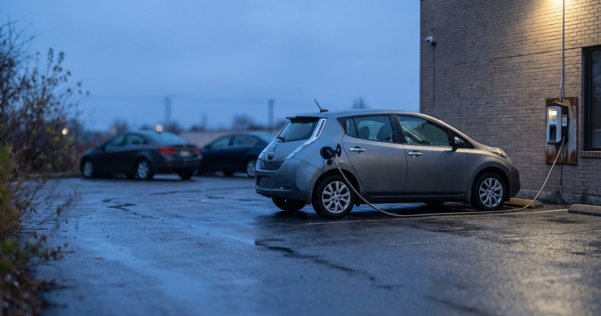 A compact electric hatchback plugged into a wall-mounted charger in a small rental lot at dusk