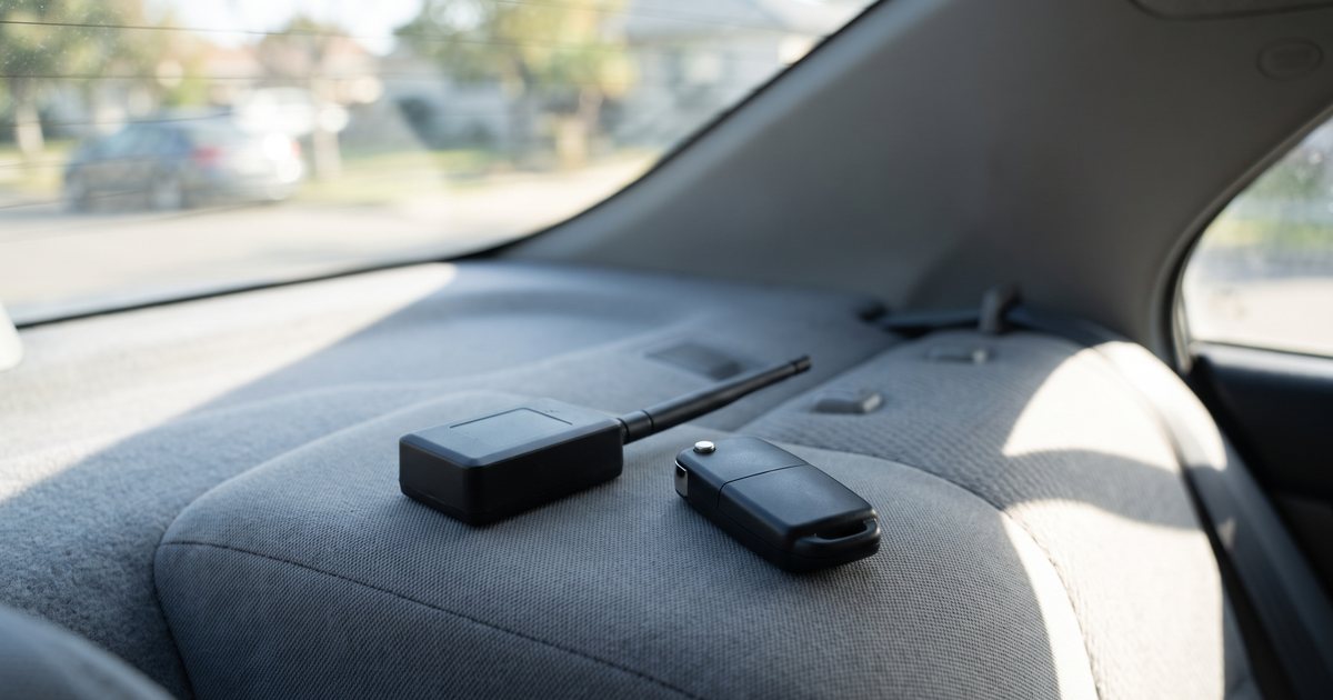 A small black GPS tracker with a short antenna and a car key fob resting on the fabric rear seat of a car