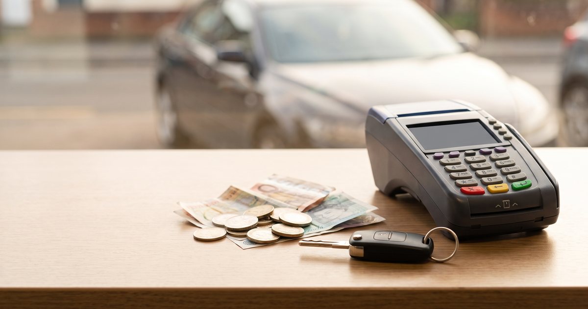 Banknotes and coins, a card terminal and a car key on a rental desk, with a car parked outside the window